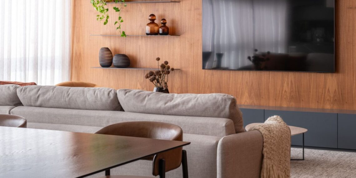 O living integrado ao home theater destaca o painel de madeira freijó, que aquece o ambiente e cria uma base visual elegante para o sofá em tom cru. Vasos artesanais, plantas naturais e prateleiras metálicas completam a composição, reforçando o equilíbrio entre aconchego e modernidade. | Imagem: Raul Fonseca (@raulfonseca)