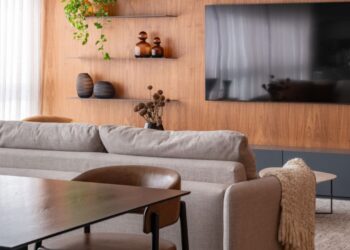 O living integrado ao home theater destaca o painel de madeira freijó, que aquece o ambiente e cria uma base visual elegante para o sofá em tom cru. Vasos artesanais, plantas naturais e prateleiras metálicas completam a composição, reforçando o equilíbrio entre aconchego e modernidade. | Imagem: Raul Fonseca (@raulfonseca)