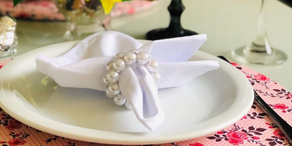 Porta-guardanapo com acabamento em pérolas brancas compõe uma mesa posta delicada e charmosa. O guardanapo de tecido branco ganha destaque sobre o prato de cerâmica e sousplat floral, criando um visual elegante e acolhedor. | Imagem: requinte_arranjo