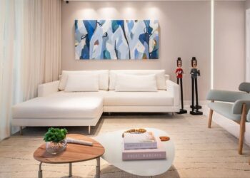 A sala ganha um ar de leveza com o sofá branco em formato lounge, que dialoga com a paleta suave das paredes e com a obra abstrata em tons de azul. As mesas de centro, em formas orgânicas, criam um ponto de encontro acolhedor, enquanto os bonecos escultóricos dão personalidade e charme ao ambiente. | Imagem: gabipontualarquitetura